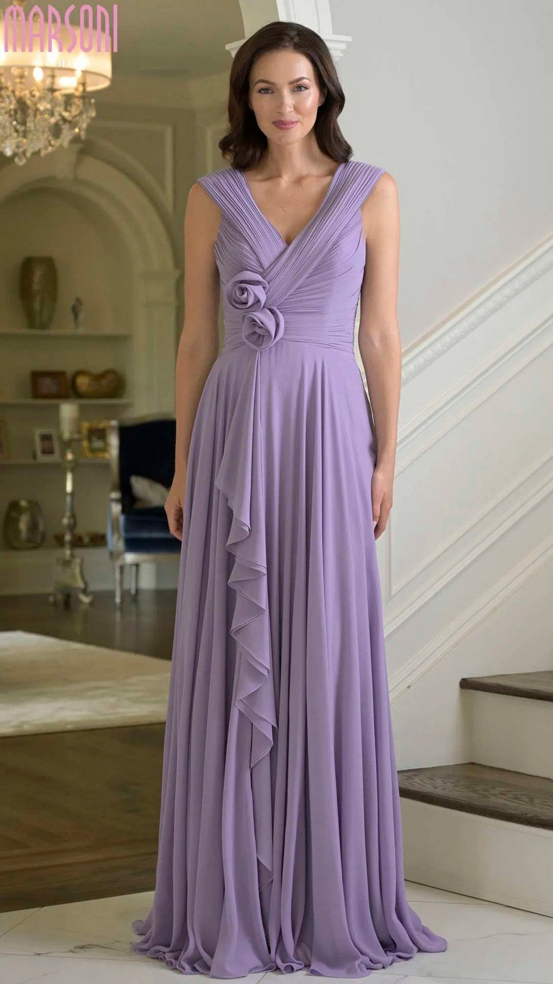 A woman stands indoors wearing the Marsoni M333 long formal dress in wisteria, featuring a sleeveless chiffon design with a v-neckline, ruffle detail, and floral embellishment at the waist.